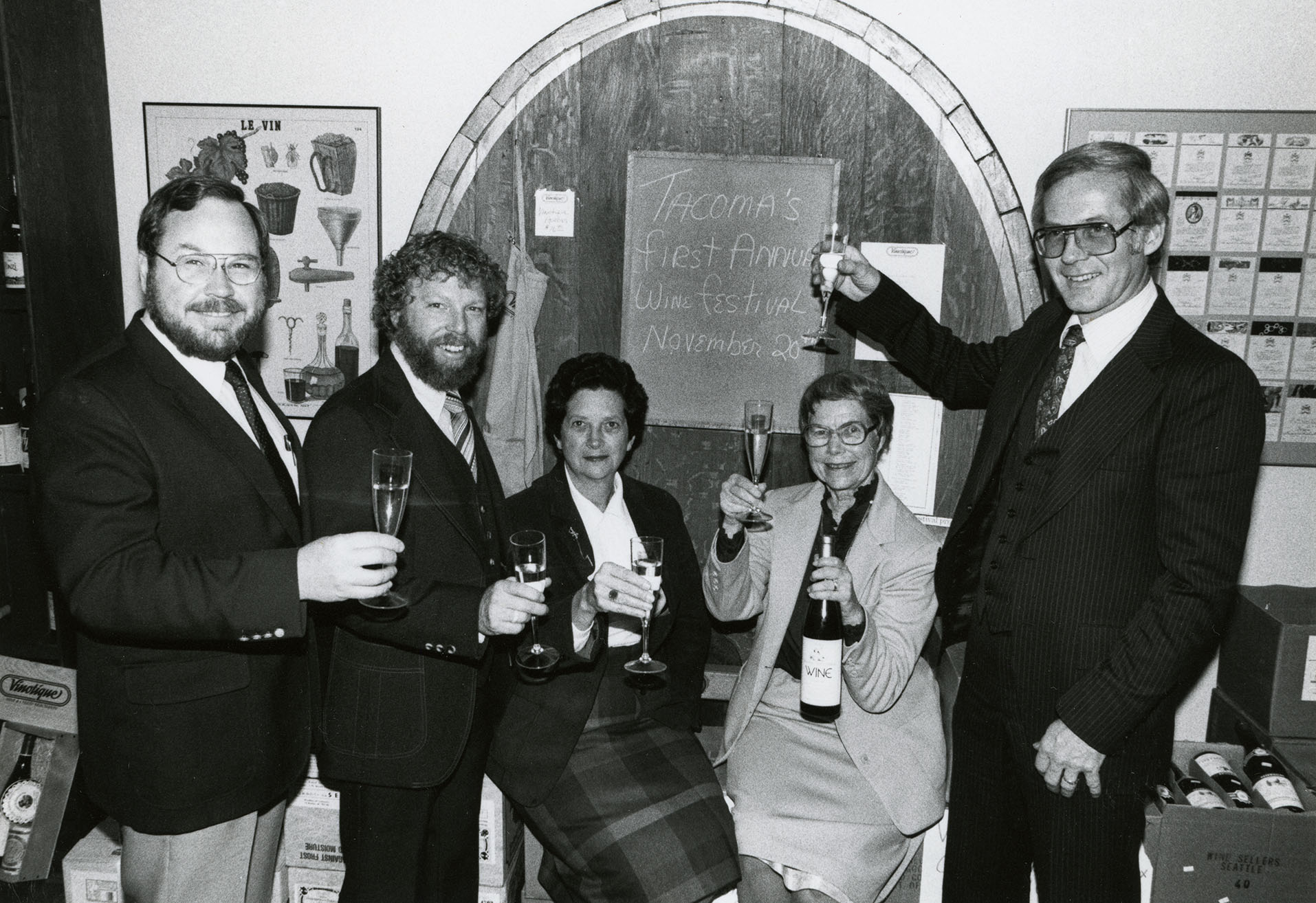Five people holding wine glasses in front of sign "Tacoma's First Annual Wine Festival November 20th"
